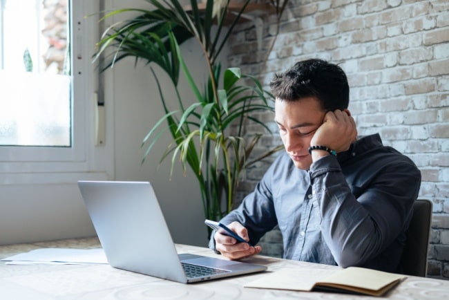 Ung man sitter ensam vid sin dator på jobbet och tittar sorgset på sin telefon.